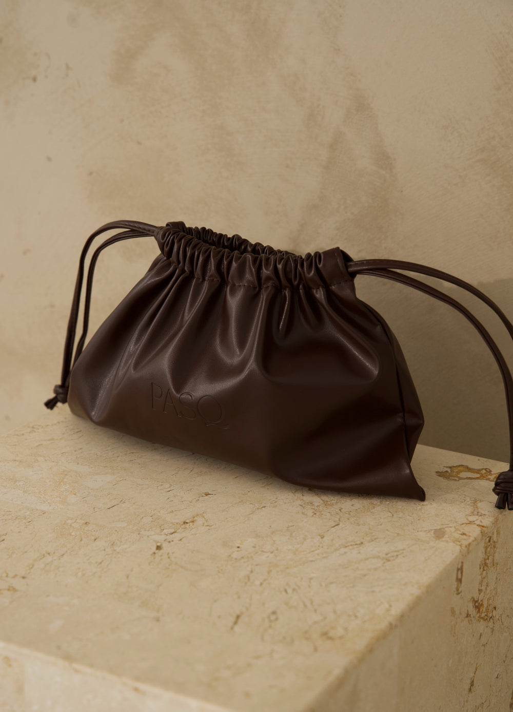 Brown leather PASQ Essentials Pouch with drawstring closure on a travertine ledge