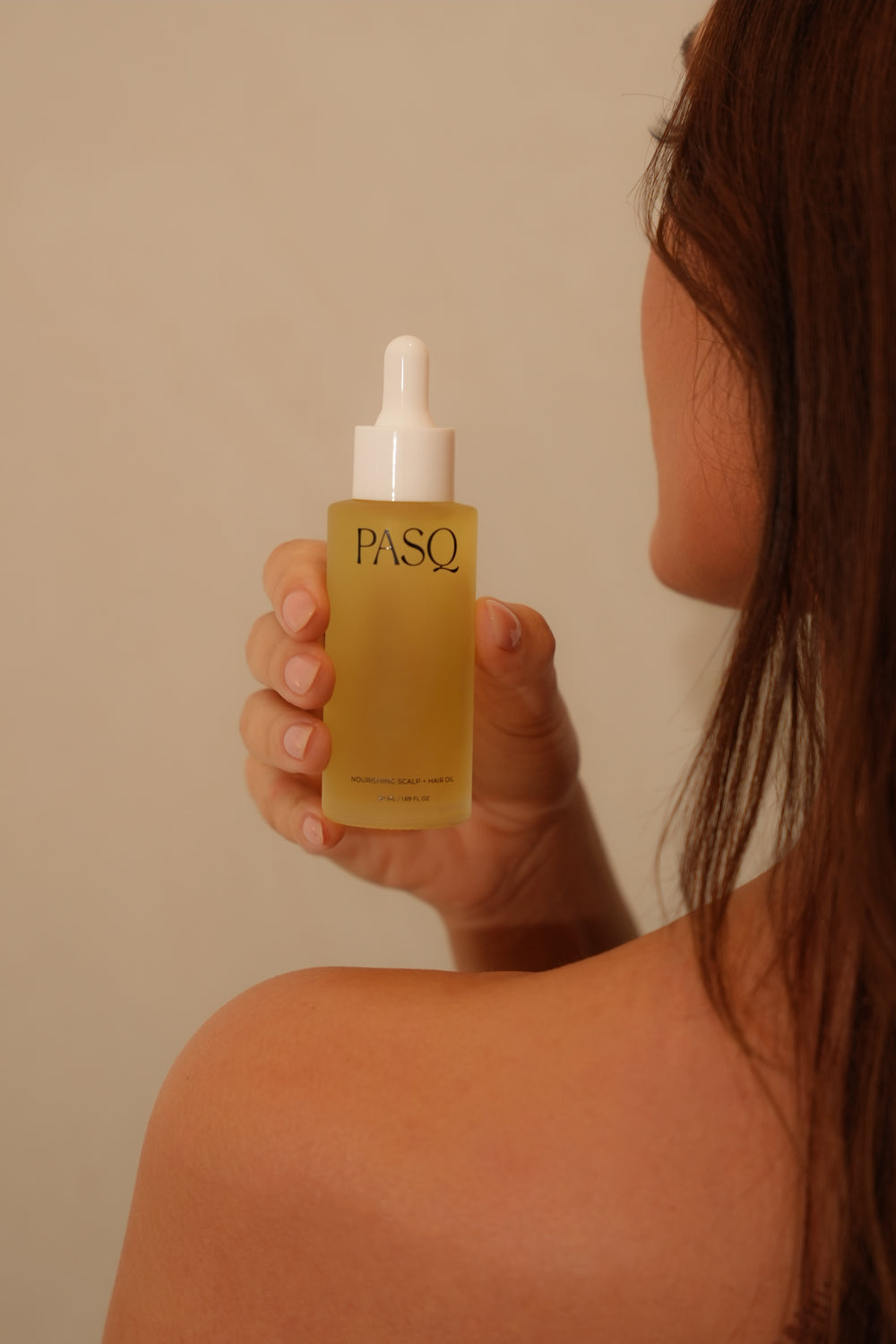 Woman seen from behind holding the amber glass PASQ Nourishing Scalp and Hair Oil dropper bottle