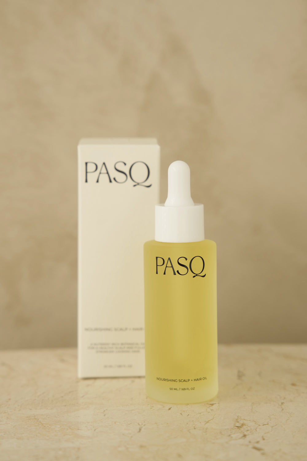 PASQ Nourishing Scalp and Hair Oil bottle beside its white carton box on a stone surface